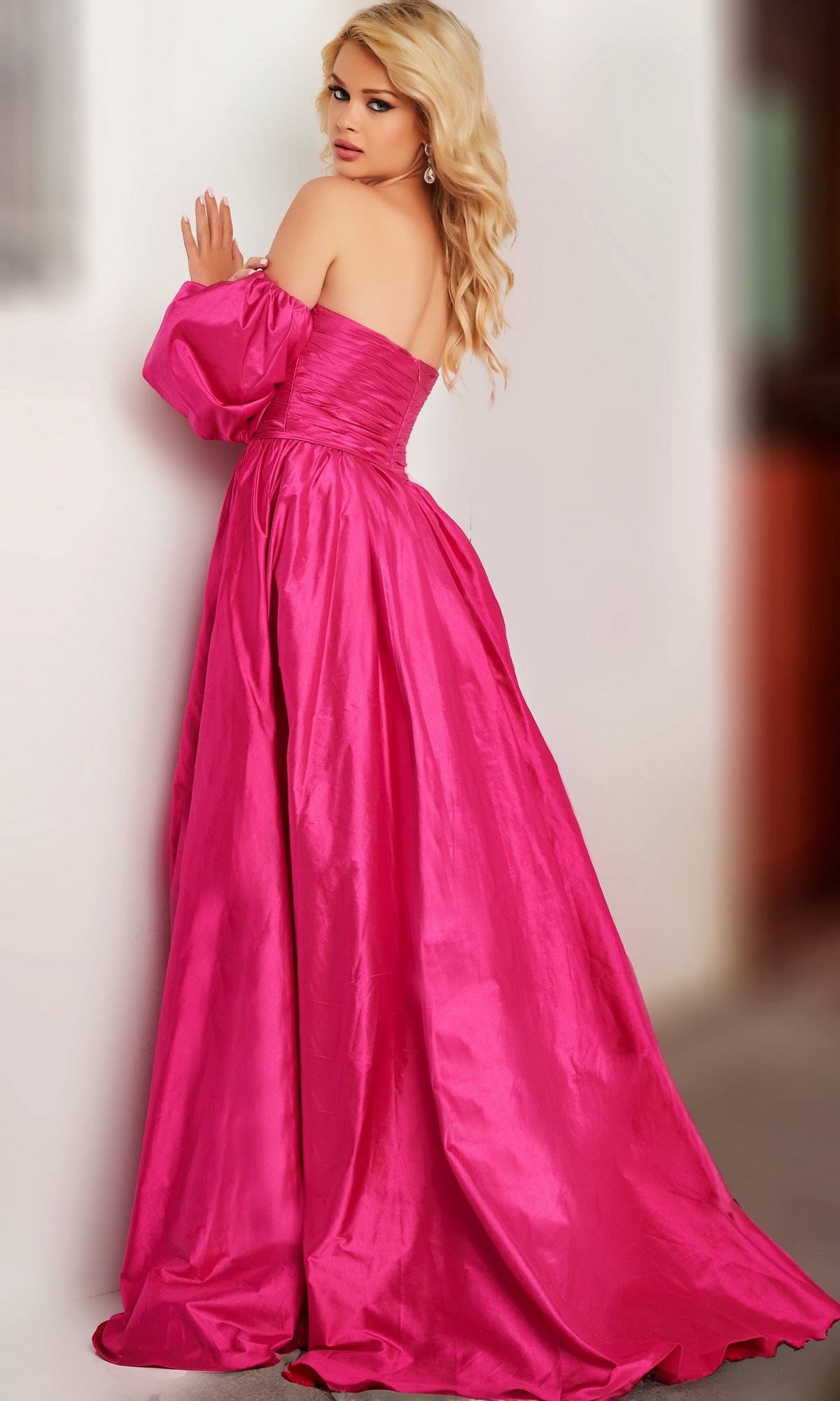 A woman with long blonde hair wears the Jovani Puff-Sleeve Long Prom Ball Gown 24099, featuring a bright pink full-length skirt, as she stands indoors and looks over her shoulder.